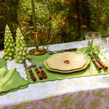 Outdoor table setting with floral patterned plates on green placemats, silverware with brown handles, two green pine tree-shaped candles, and a brass candle holder with a green candle.
