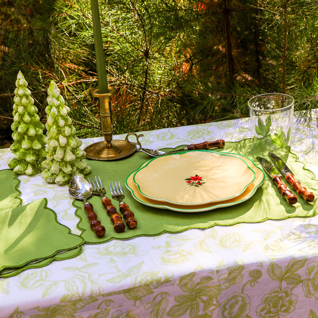 Outdoor table setting with floral patterned plates on green placemats, silverware with brown handles, two green pine tree-shaped candles, and a brass candle holder with a green candle.