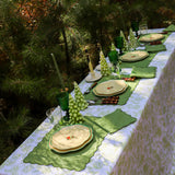 Outdoor dining table with green scalloped placemats, cream plates with floral centers, green goblets, and pinecone-shaped green candles, surrounded by lush pine trees.