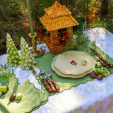 Outdoor table setting with green and white floral plates, bamboo-handled cutlery, green napkins shaped like leaves, decorative frosted green pine tree candles, and a wicker lantern with red berries.