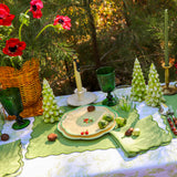 Outdoor table set with green tree-shaped candles, green glassware, floral plates, and a basket of red poppies, surrounded by natural greenery.