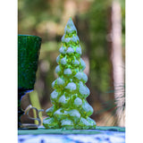 Green translucent glass pine tree sculpture with rounded needle details, standing upright on a surface with blurred outdoor woodland background and a textured green glass goblet beside it.