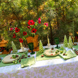 Outdoor dining table set with green and cream plates, green goblets, green leaf-shaped placemats, and a basket of red flowers under pine trees.