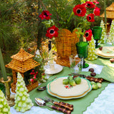 Table set with green and cream plates, bamboo-handled cutlery, green glassware, and centerpiece featuring woven wicker vase with red flowers and pine sprigs