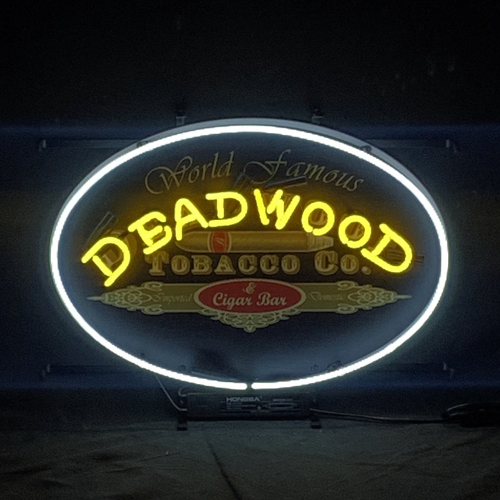 Oval neon sign with bright yellow 'DEADWOOD' lettering and a white neon border encircling 'World Famous Tobacco Co.' and 'Cigar Bar' text on dark background.