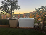 White acrylic cold plunge tub next to a beige chiller unit on a wooden deck overlooking a hilly landscape with trees and houses at sunset.