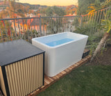 White rectangular cold plunge tub filled with water on a wooden slatted platform beside a black and beige chiller unit, set outdoors near a railing and greenery at sunset.