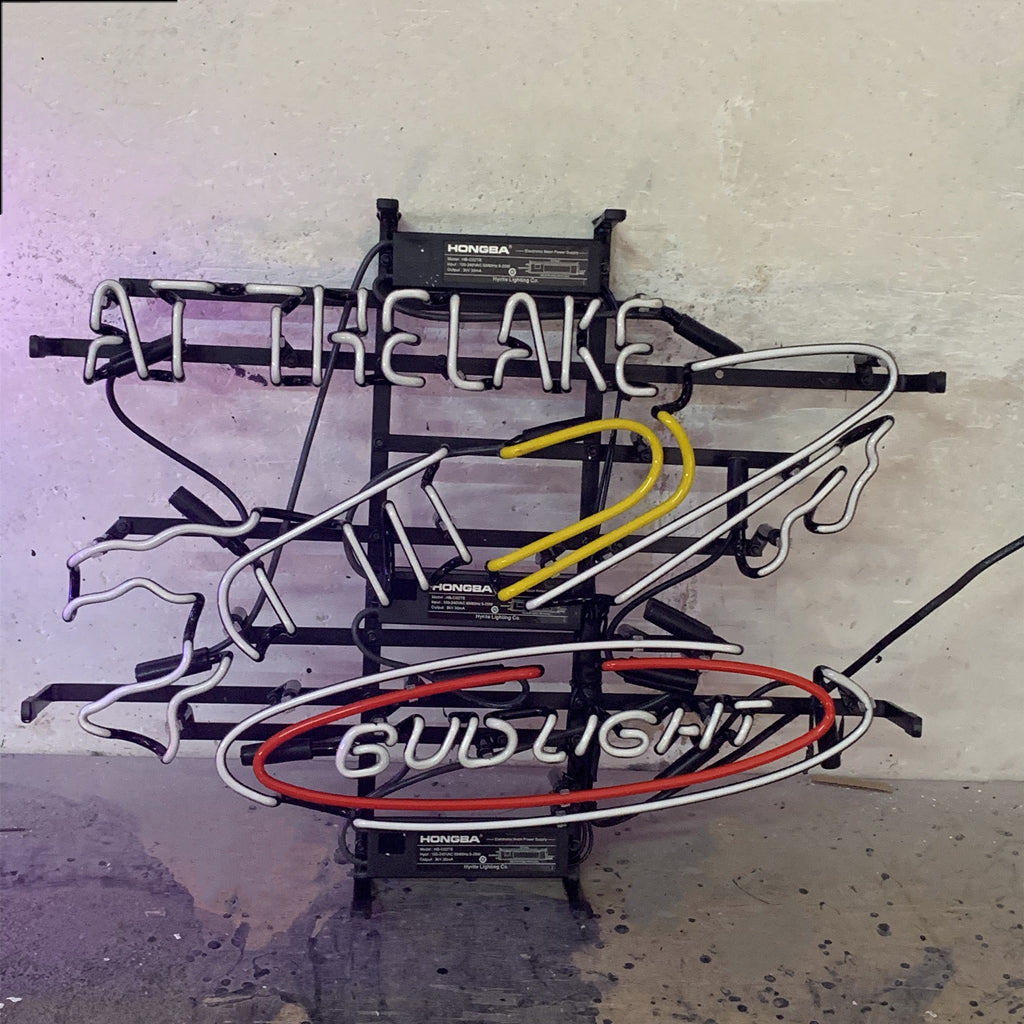 Horizontal neon sign shaped like a yacht with white, yellow, and red tubes forming the yacht and an oval around the text 'BUD LIGHT', with 'AT THE LAKE' written above in white neon tubes.