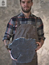 Man in plaid shirt and brown apron holding a large, weathered log cross-section with visible growth rings and cracks against a textured gray background.