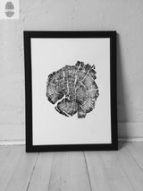 Black and white print of a tree cross-section with detailed growth rings and cracks, framed in a simple black frame leaning against a white textured wall on a light wooden floor.