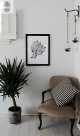 Black-framed art print of a tree cross-section with cityscape details hangs on a light wall above a beige vintage chair with a black-and-white houndstooth pillow and a potted plant on the floor.