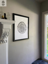 Black-framed print of an oak tree cross-section with visible rings, hanging on a beige wall near a white mantel and a window with outdoor greenery.
