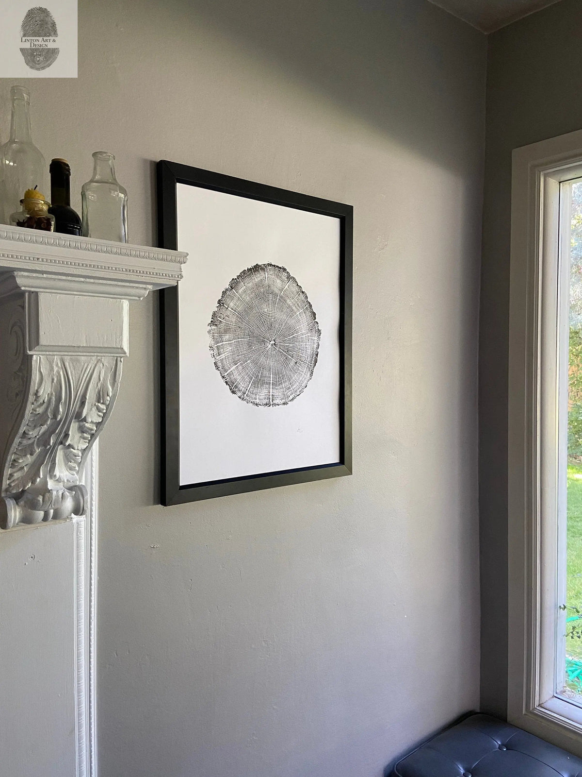 Black-framed print of an oak tree cross-section with visible rings, hanging on a beige wall near a white mantel and a window with outdoor greenery.
