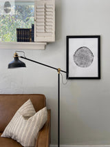 Black and white circular tree ring print in a black frame hanging on a gray wall above a brown leather sofa with two patterned pillows and next to a black adjustable floor lamp.
