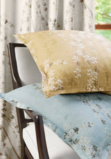 Two floral-print pillow covers, one mustard yellow and one muted blue, with white blossoms on a wooden chair against matching floral curtains.