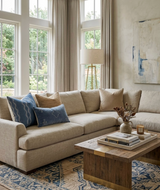 Beige sectional sofa with indigo blue Zanzibar Pillow Covers featuring white wave patterns, set in a bright living room with wooden coffee table, floor lamp, and abstract wall art.