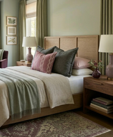 Bed with natural wood headboard dressed in gray, pink, and patterned pillows with light beige and sage green throws, flanked by wooden nightstands with mauve lamps.