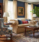 Living room with beige rolled-arm sofa dressed with red Zanzibar pillow and blue accent pillows, a floral upholstered armchair, dark wood coffee table, and navy blue walls.