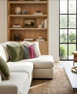 Cream sectional sofa with green velvet and berry-striped accent pillows, positioned beside a wooden built-in shelf and large black-framed window overlooking greenery.