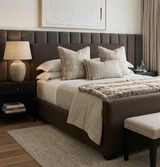Neutral bedroom with a dark soft upholstered bed featuring vertical tufted headboard panels, beige patterned and striped decorative pillows, and a light textured throw on linen bedding.