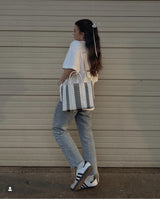 Person standing sideways against a beige corrugated wall, wearing light blue jeans, white oversized t-shirt, white sneakers with black stripes, and holding a zebra-patterned woven bag with rounded handles.