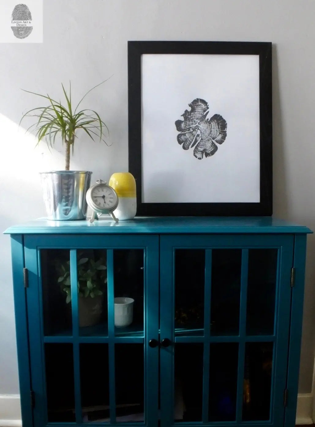 Black-framed abstract print resembling a wood cedar cross-section with fine details, displayed above a teal cabinet with glass-paneled doors and houseplants.
