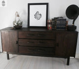 Dark wooden sideboard with two cabinets and three drawers, topped with a glass cloche containing plants, framed monochrome leaf print, stacked books, and a black vintage gramophone horn.