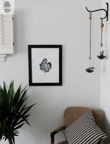 Minimalist interior with a black-framed monochrome botanical print on a light wall, a green plant in the corner, a beige chair with a black and white houndstooth pillow, and two hanging glass terrariums with rope.