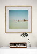 Framed photograph of people wading in shallow water under a clear blue sky, displayed on a white wall above a vintage radio and a glass vase with a single leafy stem.