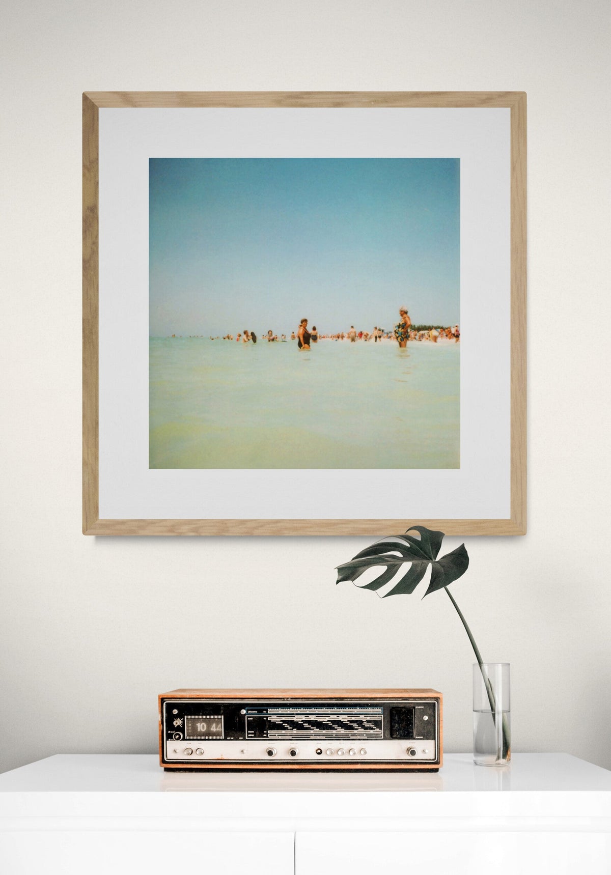 Framed photograph of people wading in shallow water under a clear blue sky, displayed on a white wall above a vintage radio and a glass vase with a single leafy stem.