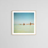 Blurred vintage-style photograph of people wading in shallow ocean water under a clear blue sky, framed with a white mat and light wooden frame.