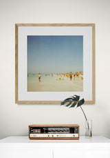 Framed fine art photograph showing a hazy beach scene with people scattered on pale sand under a soft blue sky, hanging above a vintage radio and a glass vase with a single large green leaf.