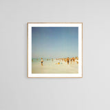 Fine art photograph showing a distant beach scene with people scattered along the shoreline under a clear blue sky, framed in light wood.