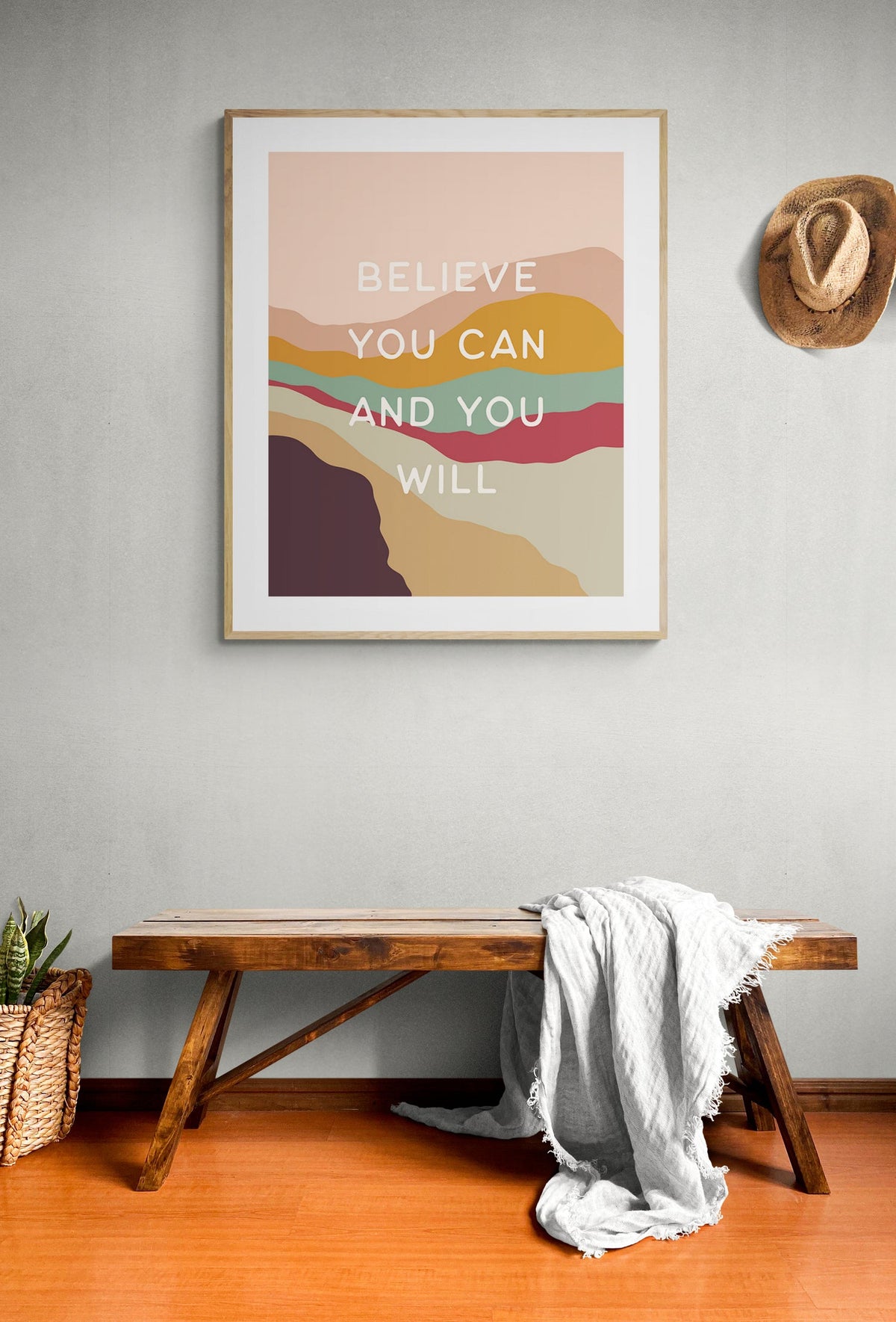 Framed modern art print with multicolored layered hills in earthy tones and text saying 'BELIEVE YOU CAN AND YOU WILL' on a light gray wall above a wooden bench with a light gray throw and a woven basket nearby.