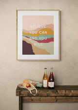 Framed modern art print with abstract layered shapes in pink, mustard, teal, and maroon hues, featuring the quote 'Believe you can and you will' in white letters.