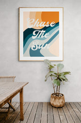 Framed modern art print with the phrase 'Chase The Sun' in white cursive over abstract waves in orange, teal, and beige, hanging on a light gray wall above a wooden floor and plant.