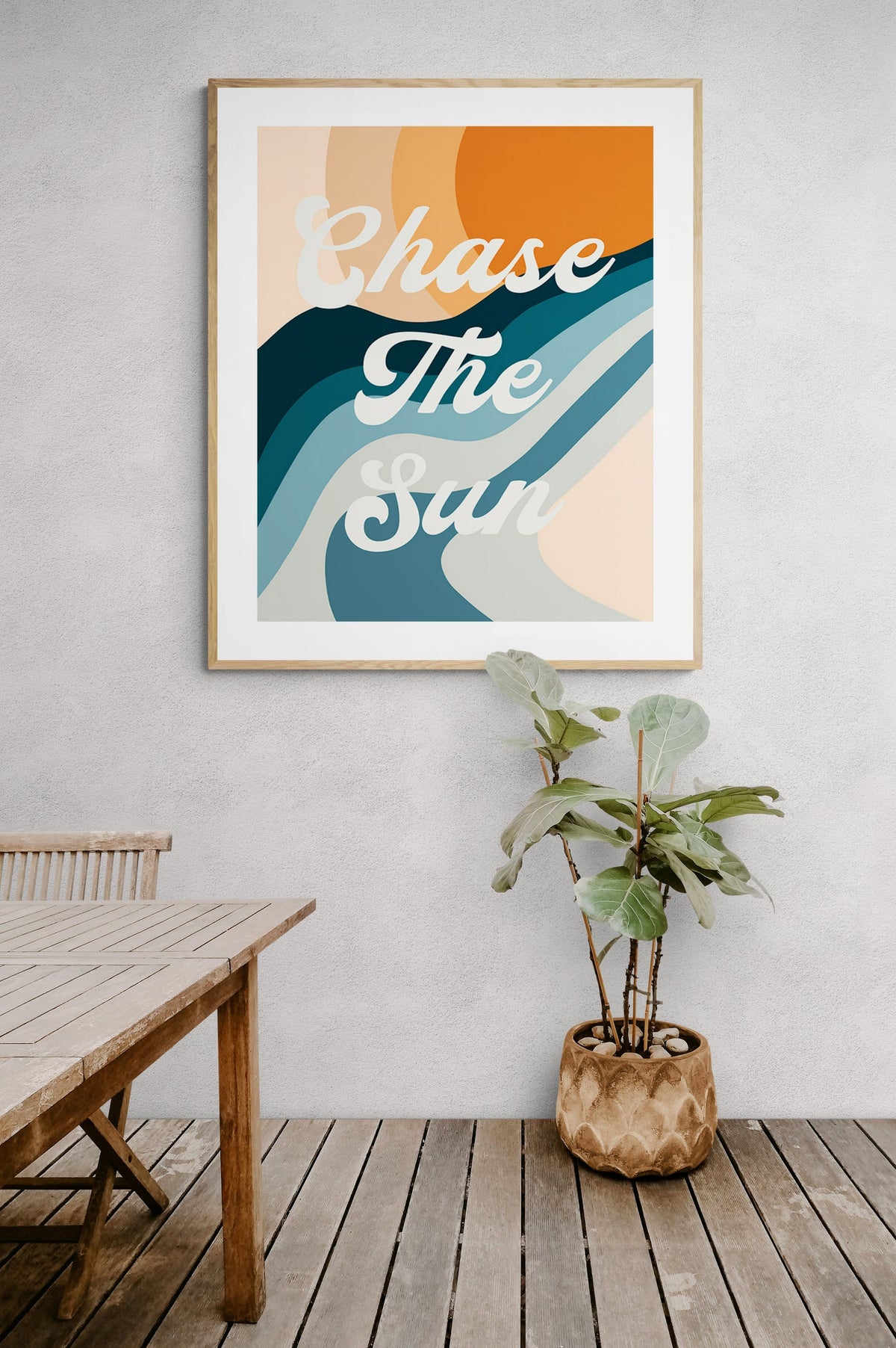 Framed modern art print with the phrase 'Chase The Sun' in white cursive over abstract waves in orange, teal, and beige, hanging on a light gray wall above a wooden floor and plant.