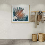 Fine art photograph of palm leaves in shades of blue and muted orange, framed in light wood and hanging on a textured beige wall.