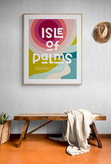 Framed modern art print with multicolored abstract shapes and text 'Isle of Palms South Carolina' hanging above a wooden bench with a light-colored blanket, next to a woven basket and cowboy hat.