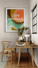 Abstract art print with organic shapes in orange, green, and brown hues featuring text 'cape HATTERAS NORTH CAROLINA' framed on a beige wall above a wooden desk with chair, plants, and window light.