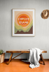 Framed modern art print with layered abstract sun and hills in warm orange, green, and brown tones, text reads 'Captiva Island Florida', hanging above a wooden bench and hat.