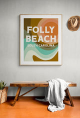Framed art print with retro-style abstract shapes in mustard, teal, coral, and cream colors, featuring bold text 'Folly Beach South Carolina' on a gray wall above a wooden bench.