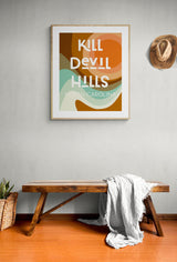 Framed modern art print with abstract earthy-toned swirls and bold white text reading 'KILL DEVIL HILLS NORTH CAROLINA' hung on a light wall above a rustic wooden bench with a draped white cloth and a straw hat hanging nearby.