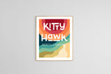 Modern art print featuring the words 'Kitty Hawk North Carolina' over layered retro color waves in red, orange, teal, green, and beige, framed on a light gray wall.