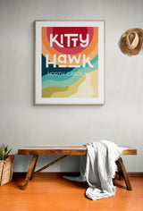 Modern art print of 'Kitty Hawk North Carolina' with colorful curved stripes in red, orange, teal, and sand tones, hanging above a rustic wooden bench with a draped white blanket and a straw hat on the wall.