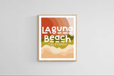 Modern art print with stylized text 'Laguna Beach California' over abstract colorful layers in red, orange, olive green, and beige tones, framed and hung on a white wall.