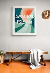 Framed modern art print with abstract teal, green, peach, and coral shapes, text reading 'Sanibel Island Florida', hanging above wooden bench with light gray throw blanket
