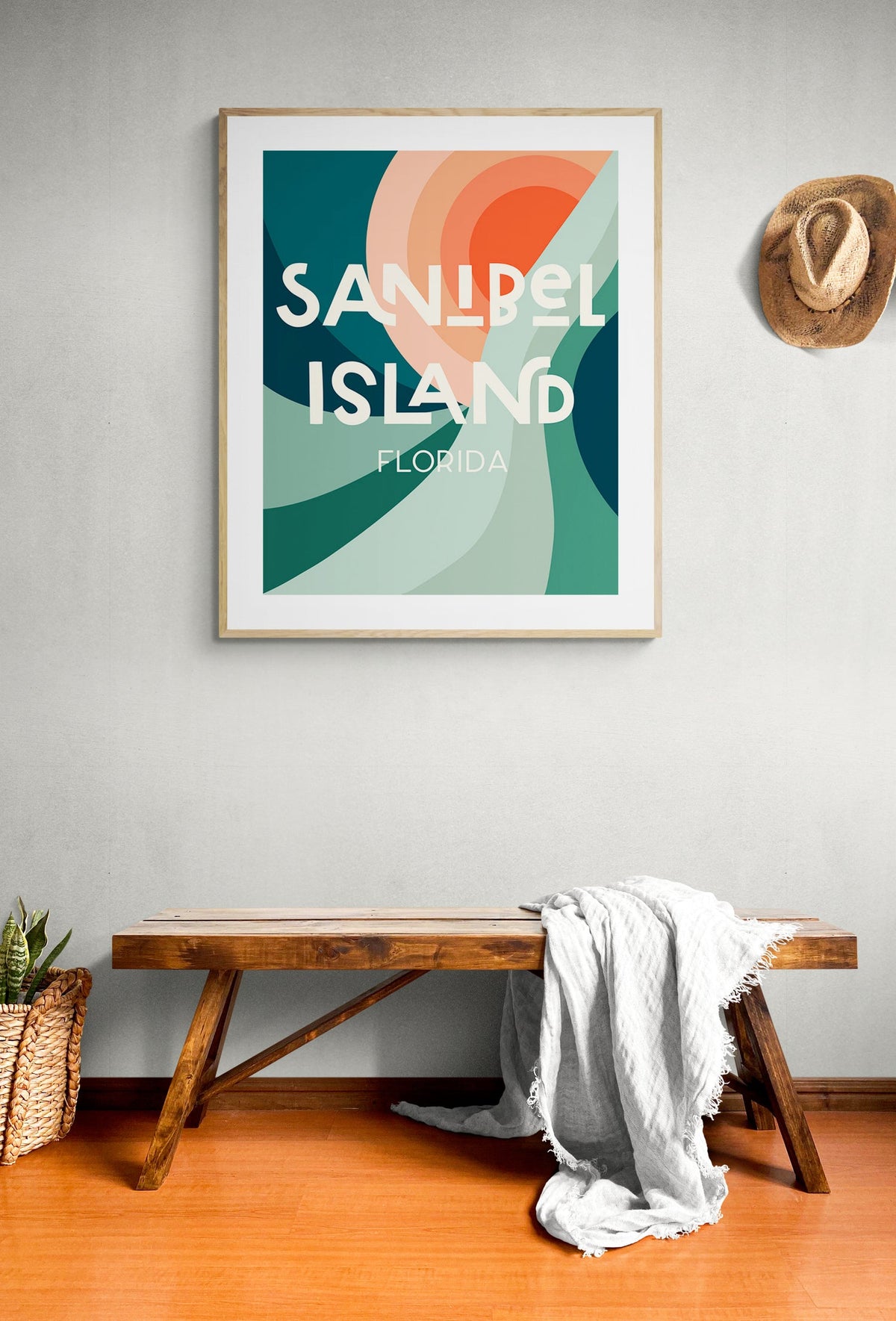 Framed modern art print with abstract teal, green, peach, and coral shapes, text reading 'Sanibel Island Florida', hanging above wooden bench with light gray throw blanket