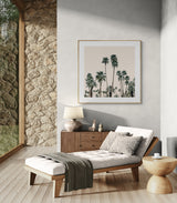 Modern living space with tufted beige chaise lounge, wooden side table, textured stone wall, and framed photograph of tall palm trees against a pale sky.