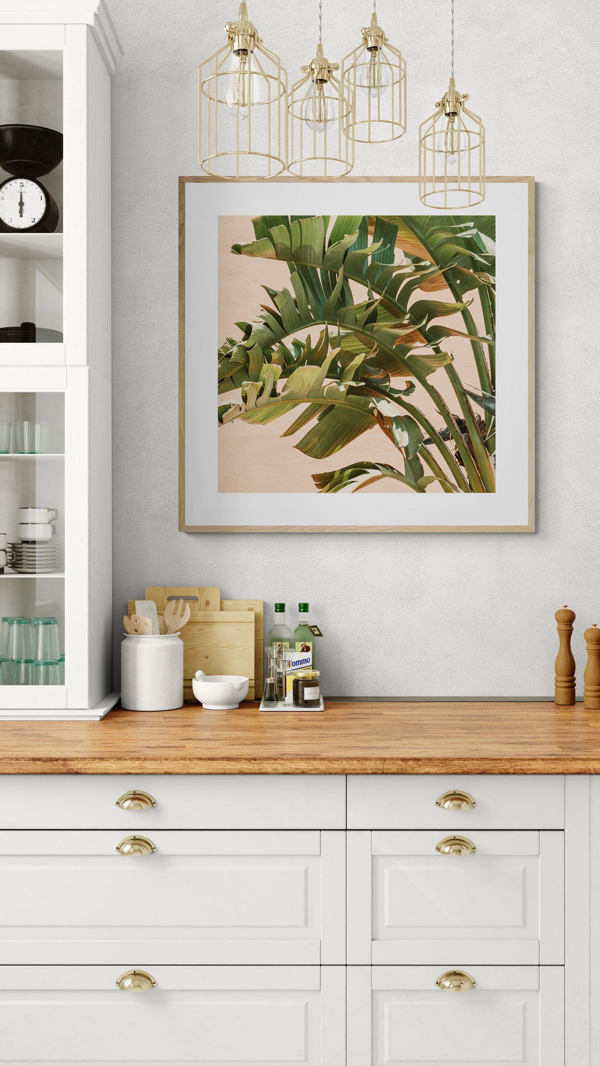 Framed photograph of green tropical leaves with peach background hanging above a white kitchen countertop with wood cutting boards, jars, and pendant lights overhead.